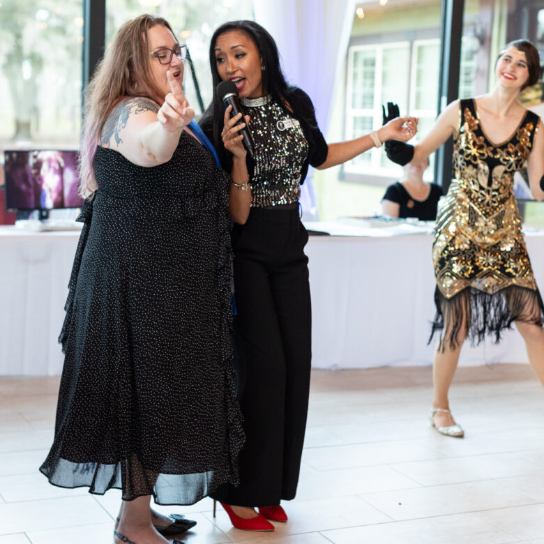 Deanna singing at an event, getting a woman involved in singing with a flapper dancer behind her.