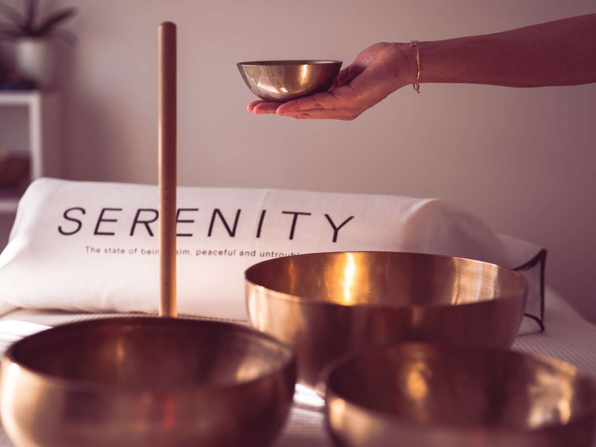 I group of tibetan singing bowls on a table with a person holding one of the bowls. In the background there is a pillow that says serenity.